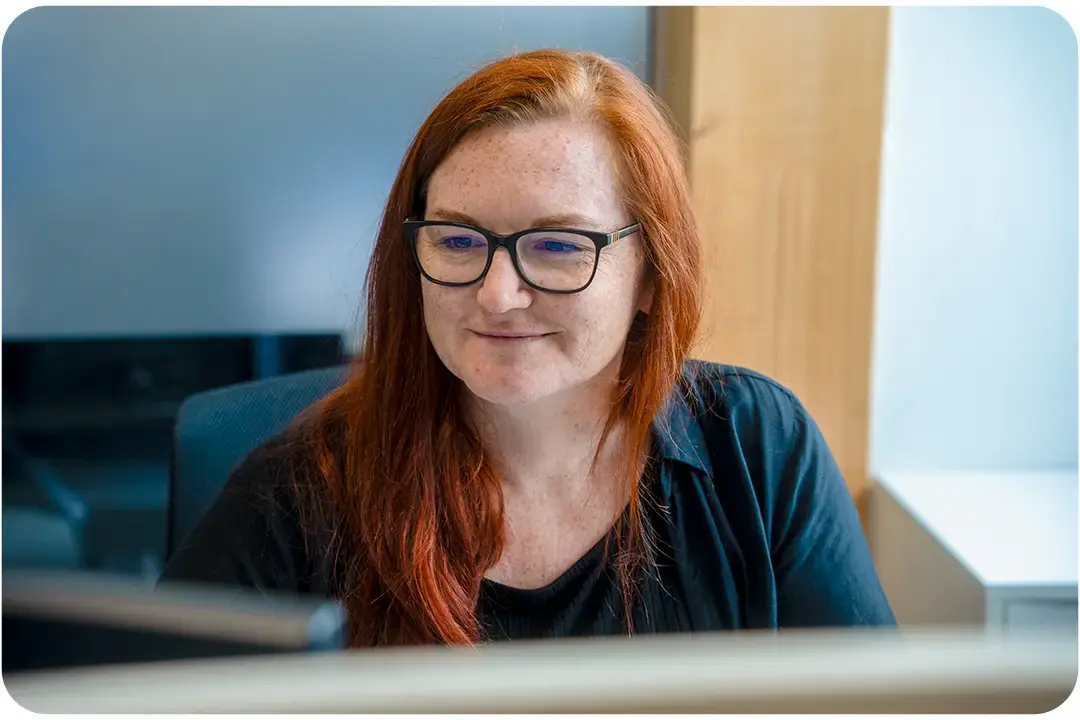 Arivo Office employee sitting smiling, diagonally in front of the camera, in front of a screen. Arivo Office employee sitting smiling, diagonally in front of the camera, in front of a screen.