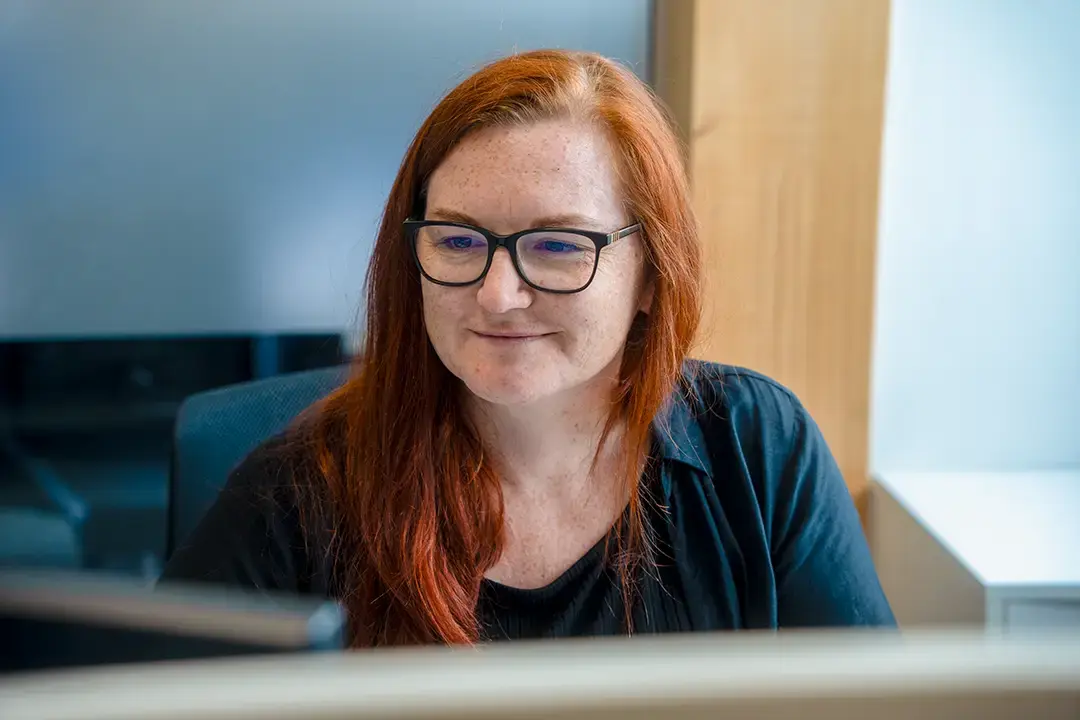 Arivo Office-Mitarbeiterin sitzt lächelnd, schräg vor der Kamera, vor einem Bildschirm. Arivo Office-Mitarbeiterin sitzt lächelnd, schräg vor der Kamera, vor einem Bildschirm.